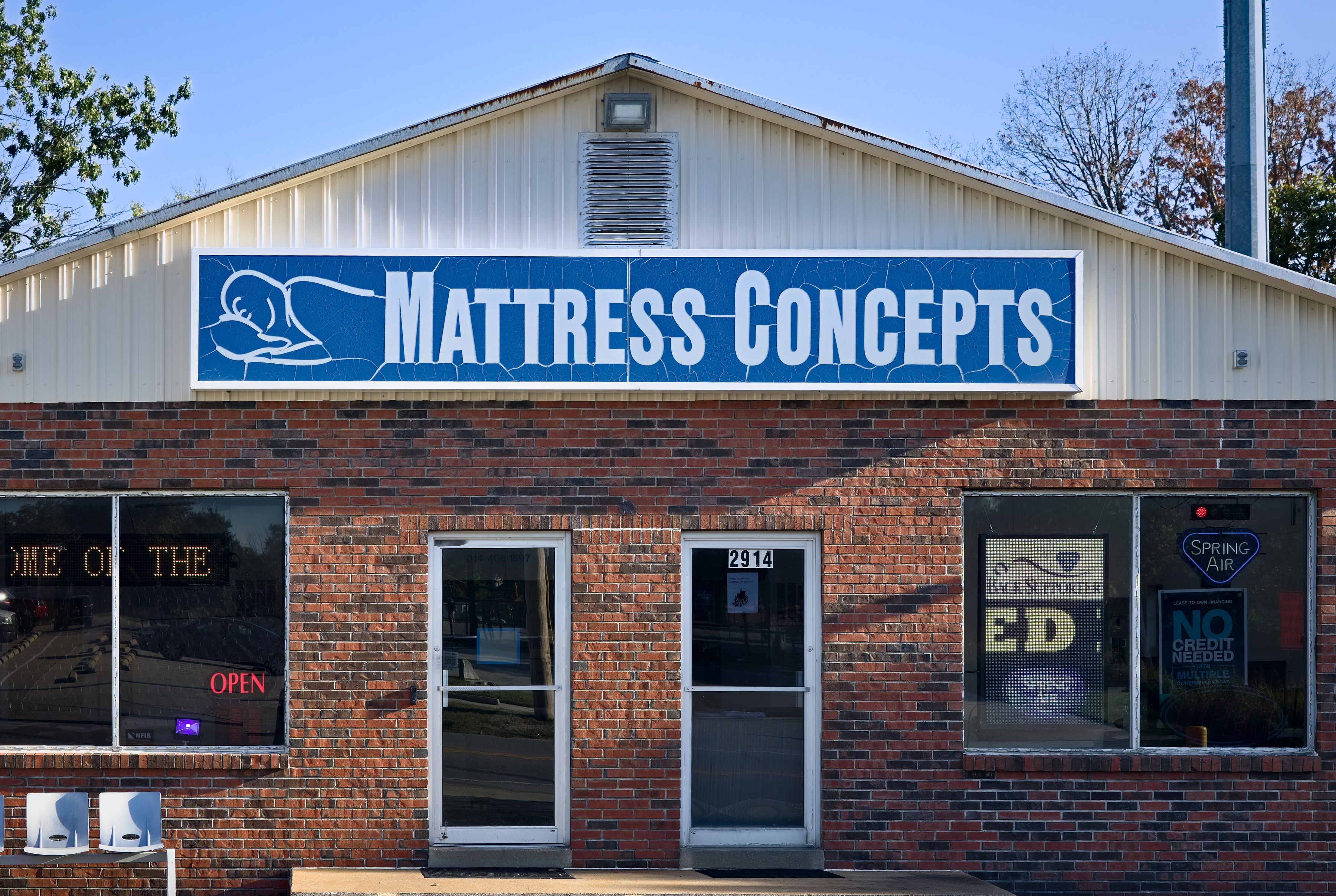 The front of Mattress Concepts in late-day sun: red brick, the blue Mattress Concepts sign and two glass doors