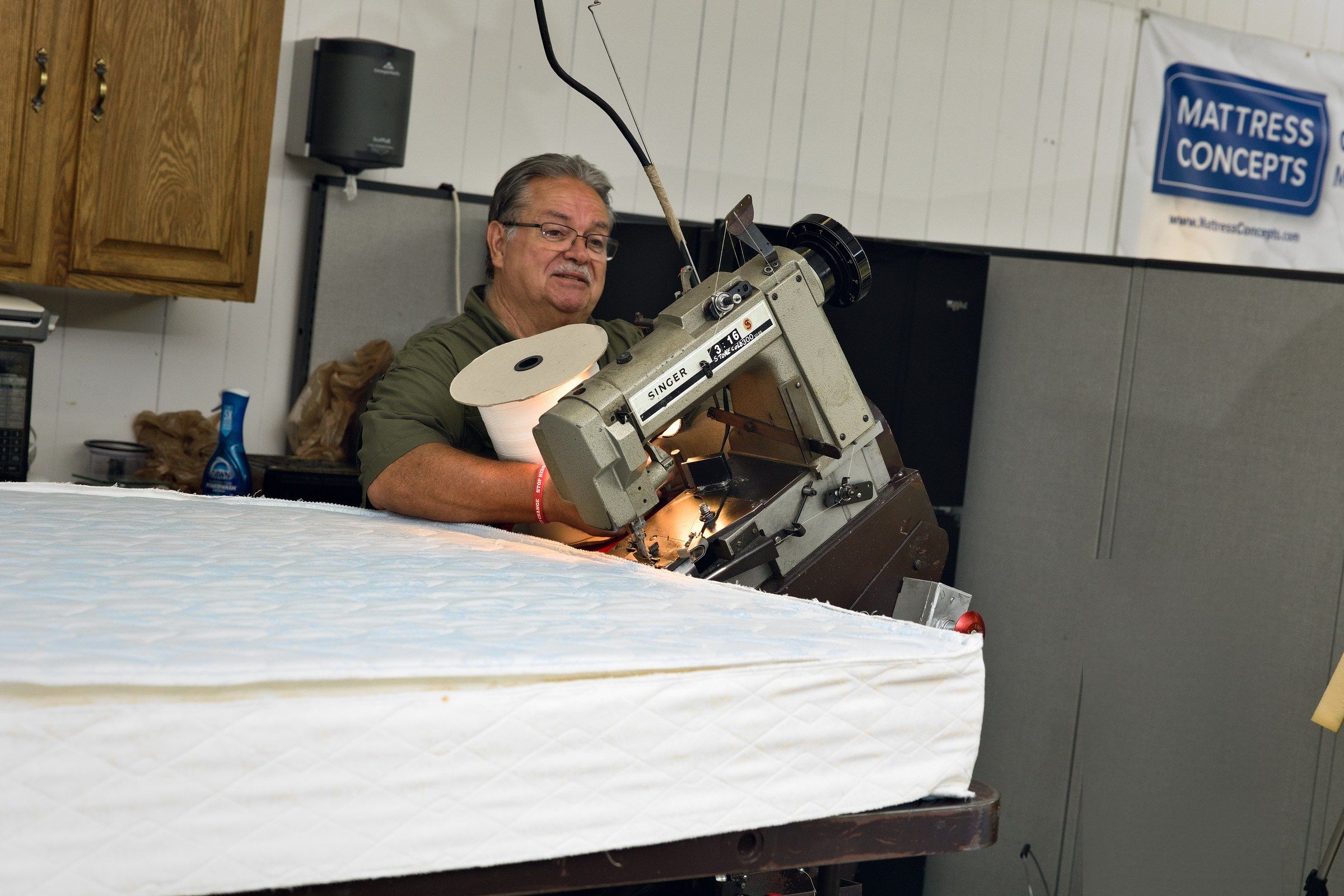 Clinton smiling behind the tape-edge machine, with the Mattress Concepts banner on the wall